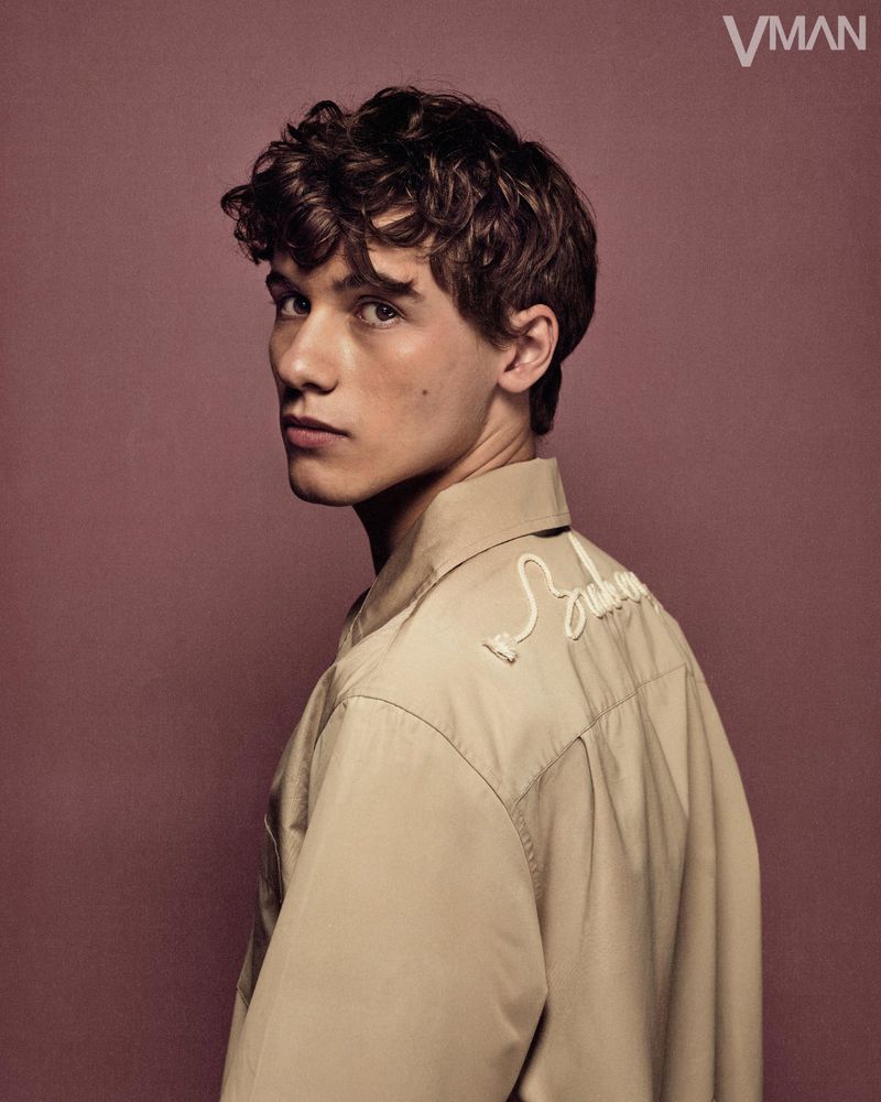 Portrait of a young adult man with curly hair looking over his shoulder, wearing a light brown shirt against a brown background.