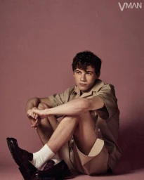 Sam Nivola seated on a solid backdrop wearing a tan short-sleeve shirt, shorts, white socks, and dark loafers.
