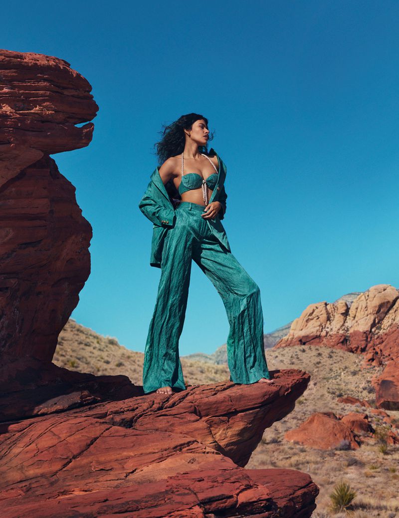 Adult female stands on red rock in turquoise two-piece outfit and jacket against a clear blue sky and desert landscape.