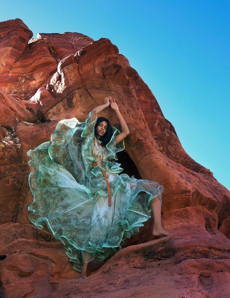 An adult female model poses in a large ruffled gown in a desert canyon with red rock formations and a bright blue sky in an editorial fashion shot.