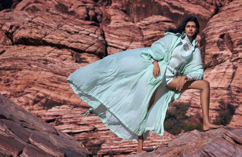 A barefoot woman in a light green coat poses on a rock formation, set against a background of layered red rock in a desert landscape.
