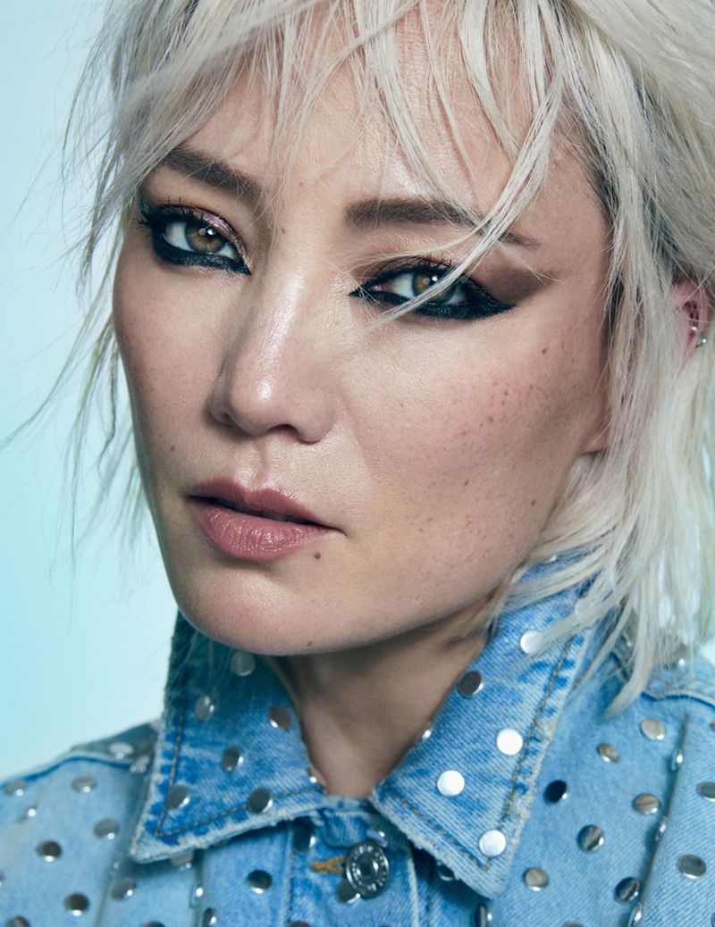 Close-up portrait of a fair-skinned woman with platinum blonde hair, wearing a studded denim jacket and heavy makeup, against a gradient background.