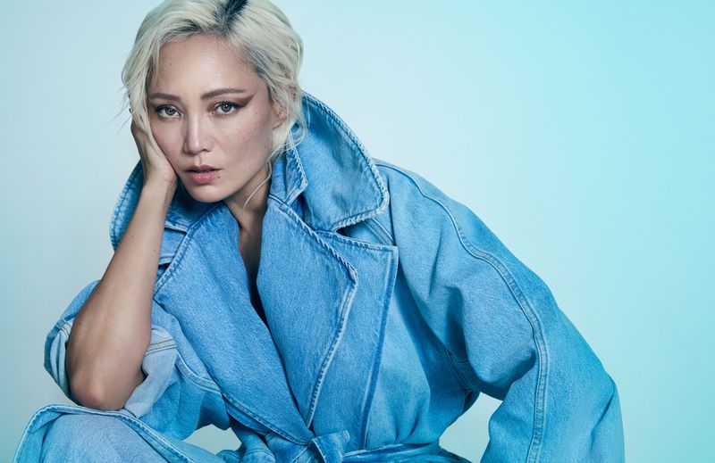Portrait of an adult female with platinum blonde hair wearing a light blue denim jacket in a studio with a teal background.