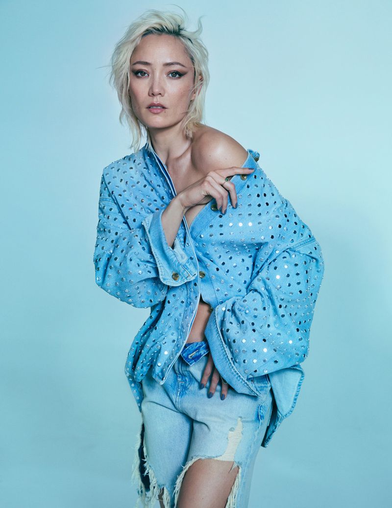 Attractive Asian woman poses in a fashionable light blue denim jacket and ripped jeans against a soft blue backdrop in a studio shot.