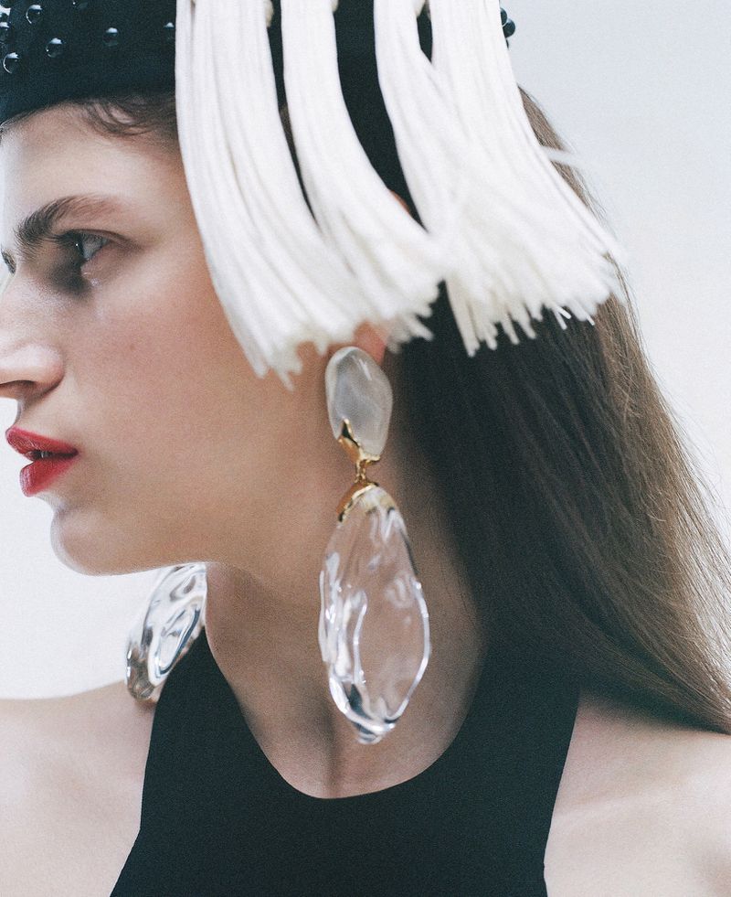 Close-up shot of a woman with white fringe headwear and large clear sculptural earrings, modelling in a minimalist setting.