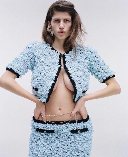 A fashionable woman poses in a light blue jacket and skirt, accessorized with a delicate pearl belt against a white background.