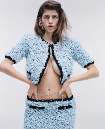 A fashionable woman poses in a light blue jacket and skirt, accessorized with a delicate pearl belt against a white background.