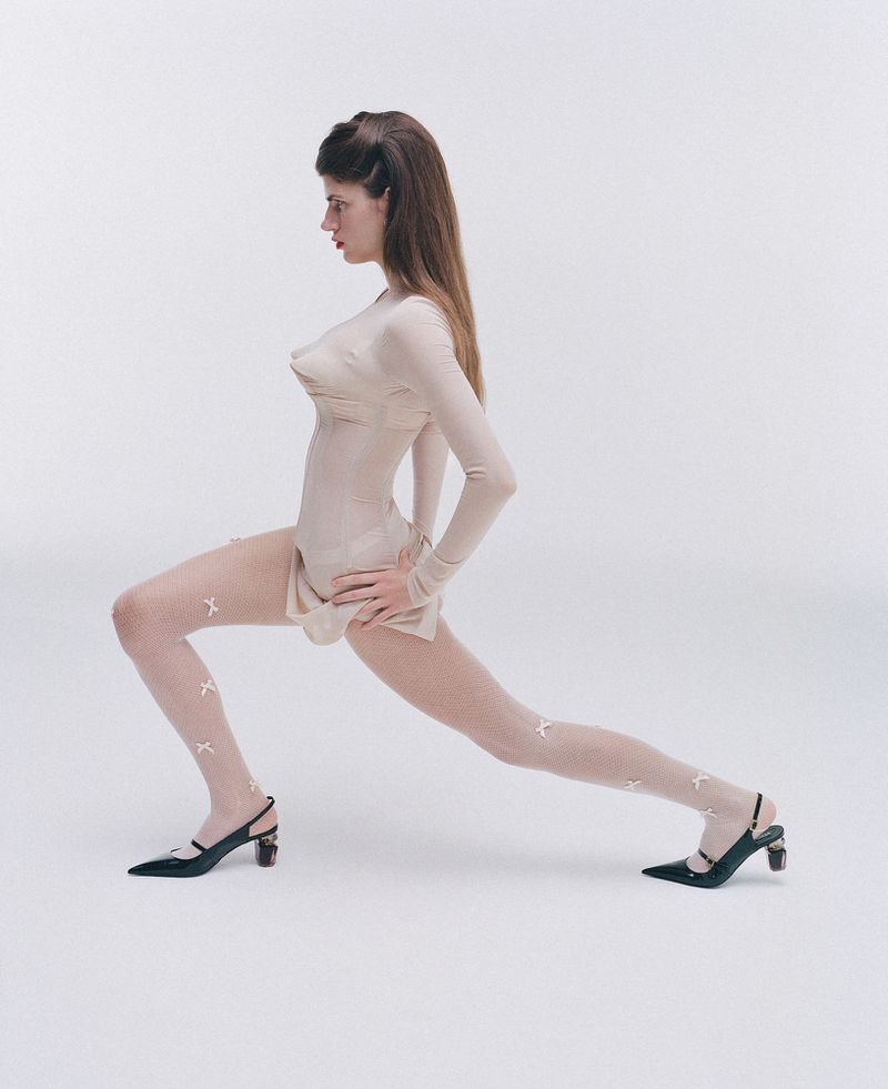 A woman in a beige bodysuit, patterned tights, and black mules in a full-body studio shot, posing in a lunge with hands on her hips.