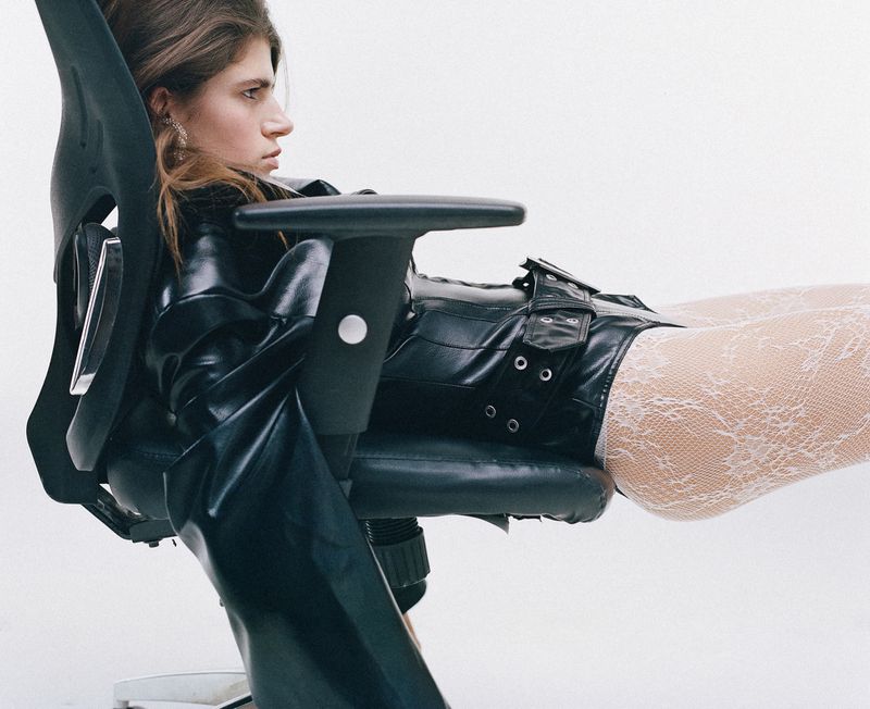 Woman in black leather outfit reclining on office chair against a bright white background. Fashion editorial with a stylish, edgy aesthetic.