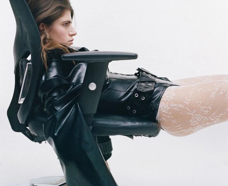 Woman in black leather outfit reclining on office chair against a bright white background. Fashion editorial with a stylish, edgy aesthetic.