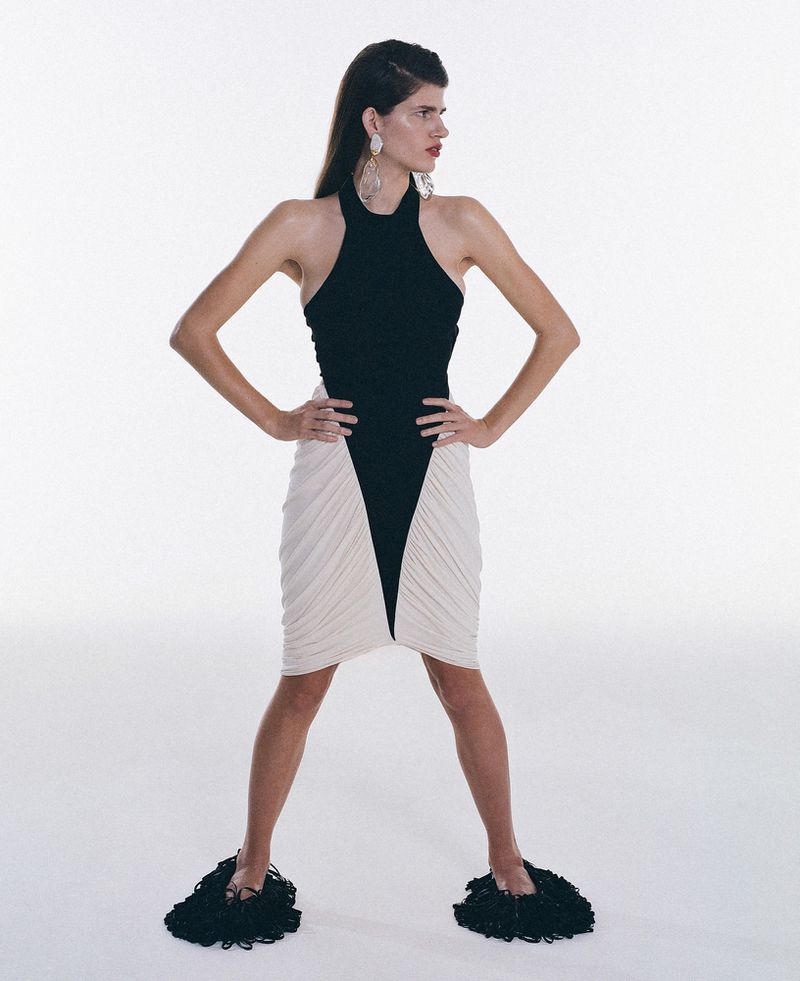 A female model poses in a black halter top and beige skirt, accessorized with large earrings and black fuzzy flats, set against a white background.