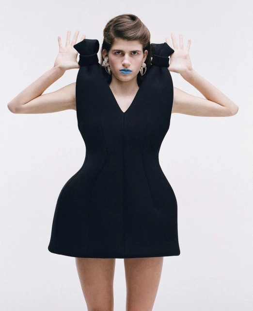 Woman in sculptural black mini dress with blue lipstick and earrings poses against a white backdrop in a fashion shot.