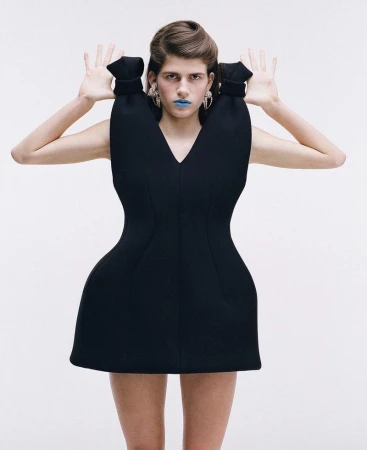 Woman in sculptural black mini dress with blue lipstick and earrings poses against a white backdrop in a fashion shot.