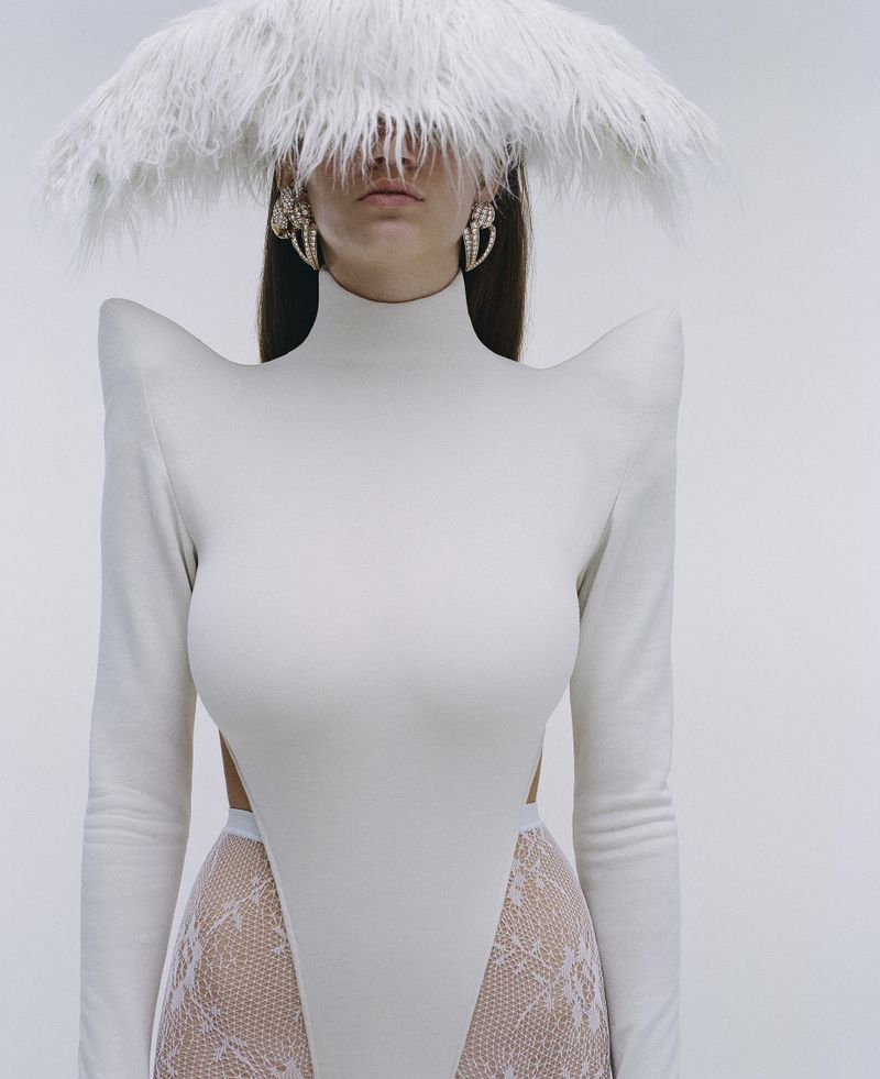 Fashionable woman in a white bodysuit with shoulder pads and lace bottoms stands against a white backdrop wearing large feather headdress.