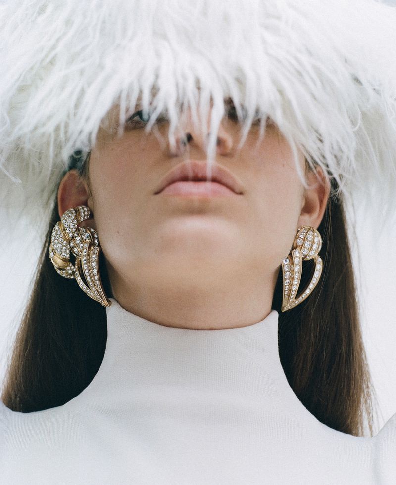 Close-up shot of an adult woman wearing large, ornate earrings and a white feathered hat with a matching top against a plain background.