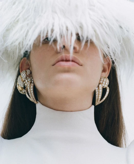 Close-up shot of an adult woman wearing large, ornate earrings and a white feathered hat with a matching top against a plain background.