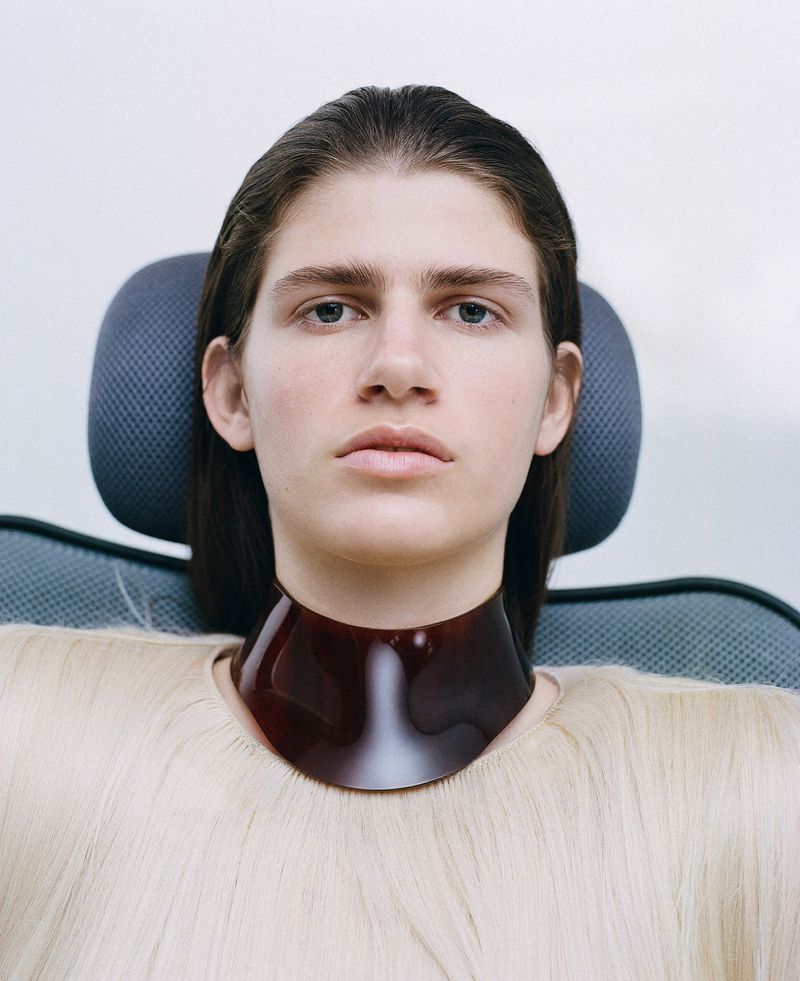 Portrait of androgynous person with clean complexion, wearing a shiny brown collar, seated in a modern chair against a light background.