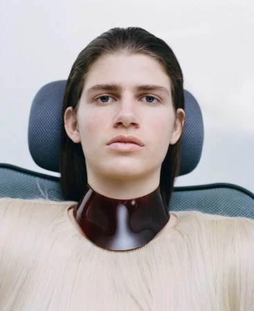 Portrait of androgynous person with clean complexion, wearing a shiny brown collar, seated in a modern chair against a light background.
