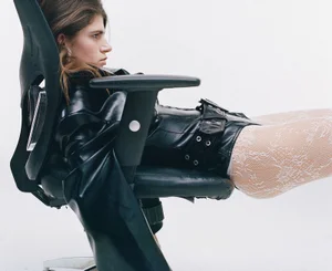 Model wearing black leather jacket and white lace tights seated horizontally in an office chair