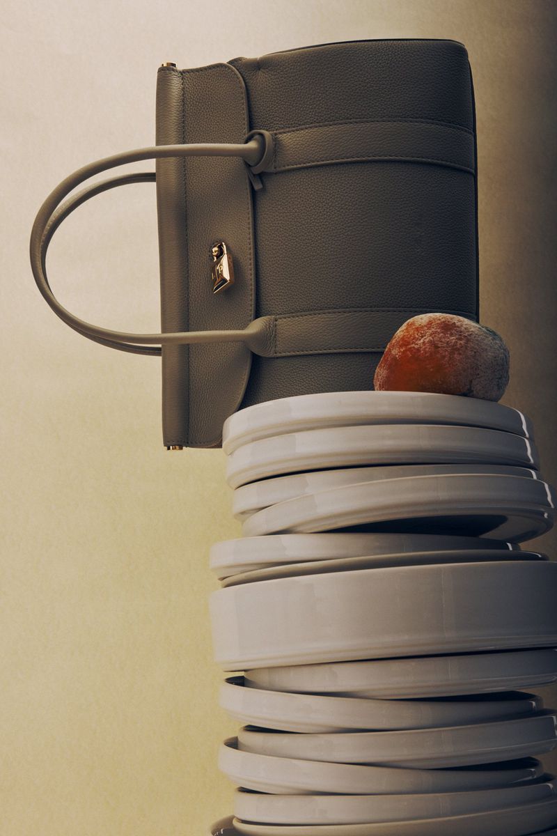 Grey leather satchel bag on a tower of white ceramic plates, with an orange resting on top, against a pale graduated background.