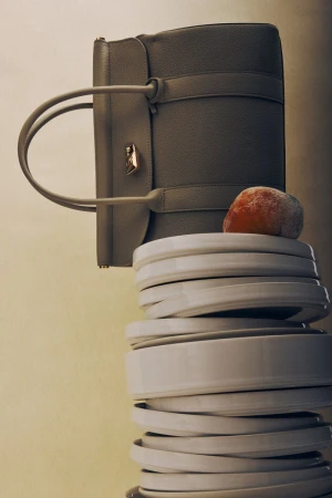 Grey leather satchel bag on a tower of white ceramic plates, with an orange resting on top, against a pale graduated background.