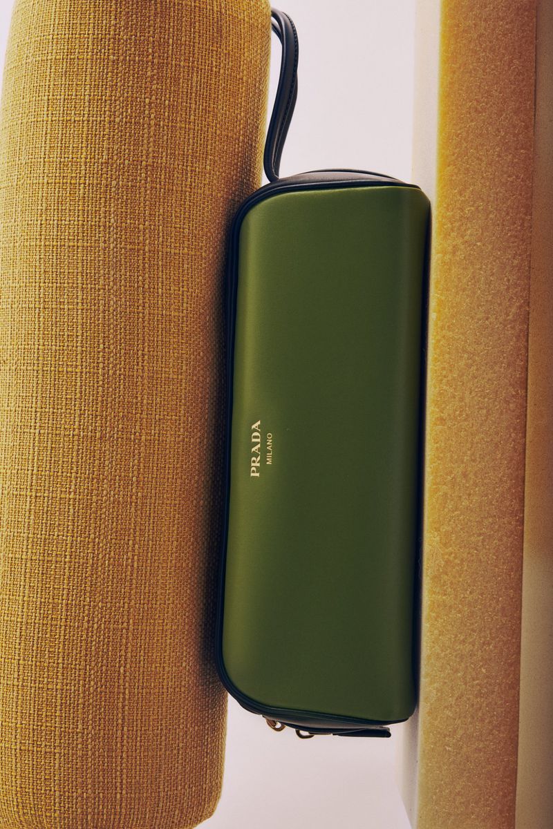 Olive green Prada Milano pouch with a black strap, nestled between textured tan and cream-colored cushions, product photography.