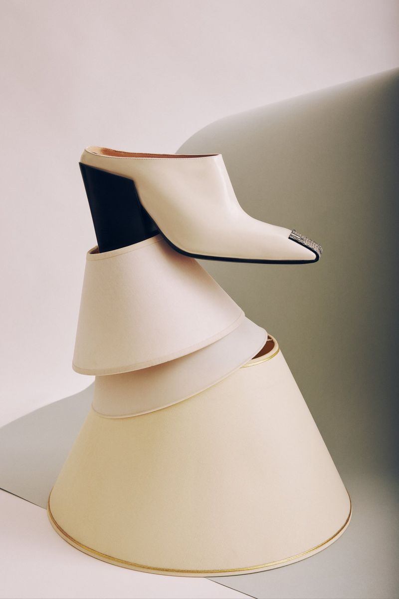Cream-colored high-heel mule is elegantly perched on a tower of lamp shades against a soft white and grey background in a fashion still life.