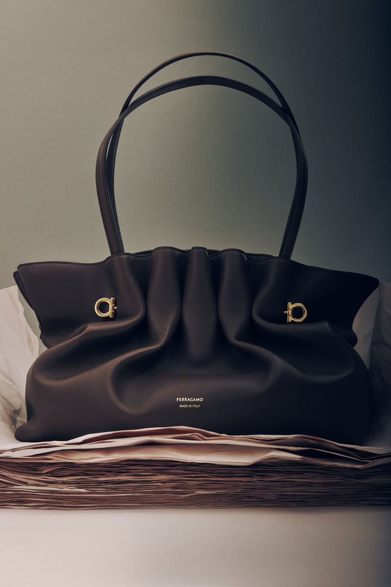 Elegant dark brown Ferragamo handbag with double handles displayed on a stack of papers against a gradient grey-green background.