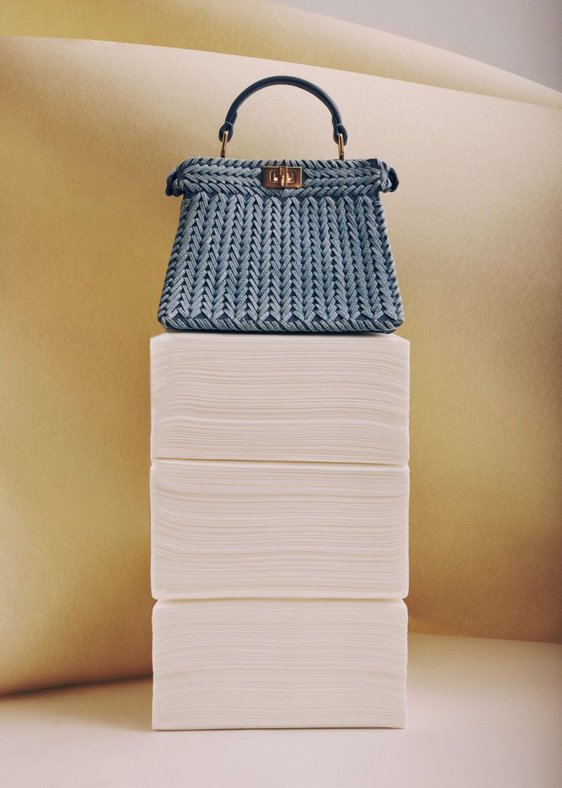 Elegant light blue woven handbag with dark blue handle sits atop a neat stack of white napkins against a pale yellow backdrop.