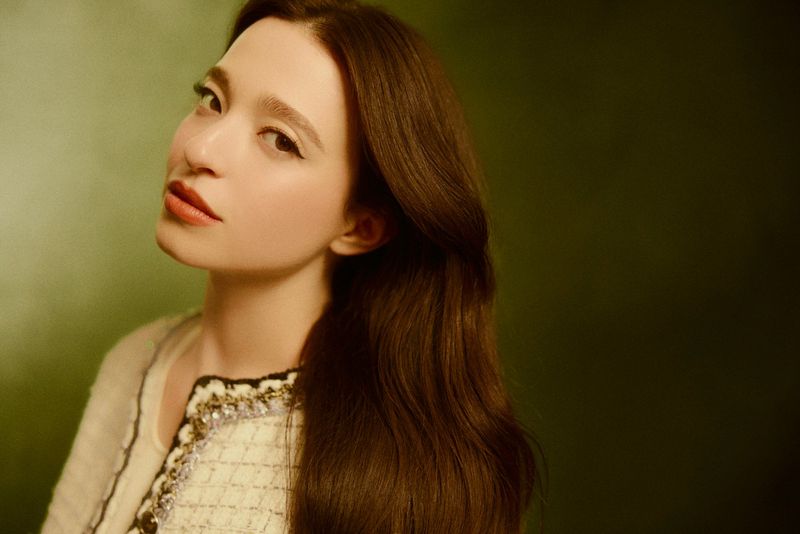 Portrait of an adult female model with long brown hair, wearing an embellished cream jacket, against a green background in a studio.