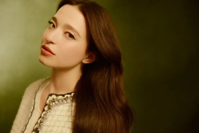 Portrait of an adult female model with long brown hair, wearing an embellished cream jacket, against a green background in a studio.