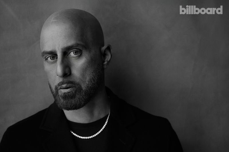 Black-and-white portrait of a bald adult male with a beard, wearing a black jacket and diamond necklace, against a gray backdrop.