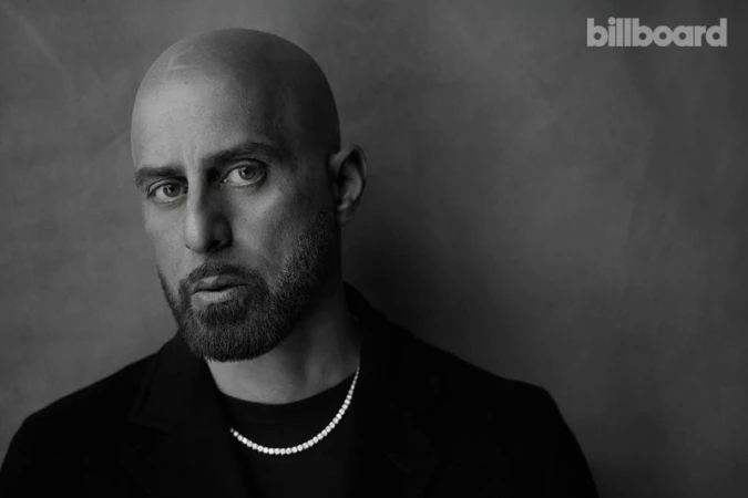 Black-and-white portrait of a bald adult male with a beard, wearing a black jacket and diamond necklace, against a gray backdrop.