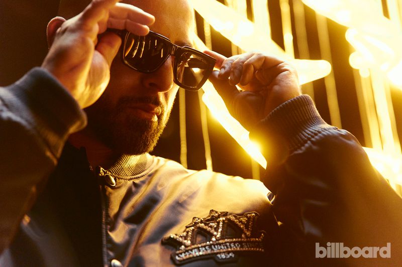 Man adjusts sunglasses in close-up, wearing a black bomber jacket with gold accents, illuminated by a bright light source.