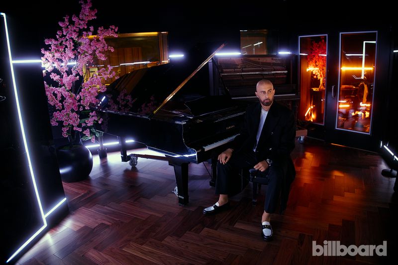 Adult male in a black suit sitting next to a glossy black grand piano with faux pink cherry blossoms in a music studio setting.