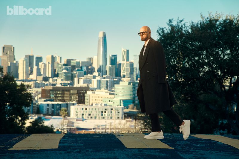 Adult male in a black overcoat walks on a crosswalk with the San Francisco skyline and blue sky in the background on a sunny day.