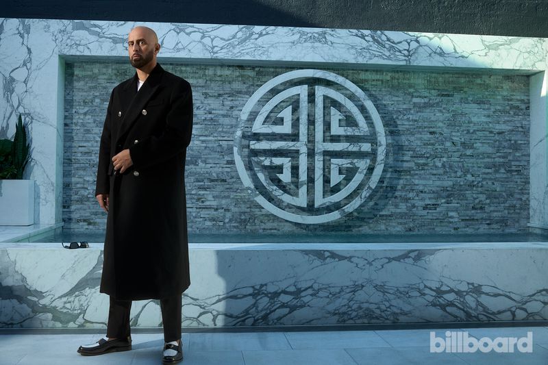 Portrait of an adult male wearing a black coat in front of a marble tile wall with decorative circle, Billboard logo in the corner.