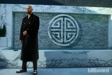 Bald man wearing a long black coat standing in front of a white marble wall with a circular logo for Billboard Magazine