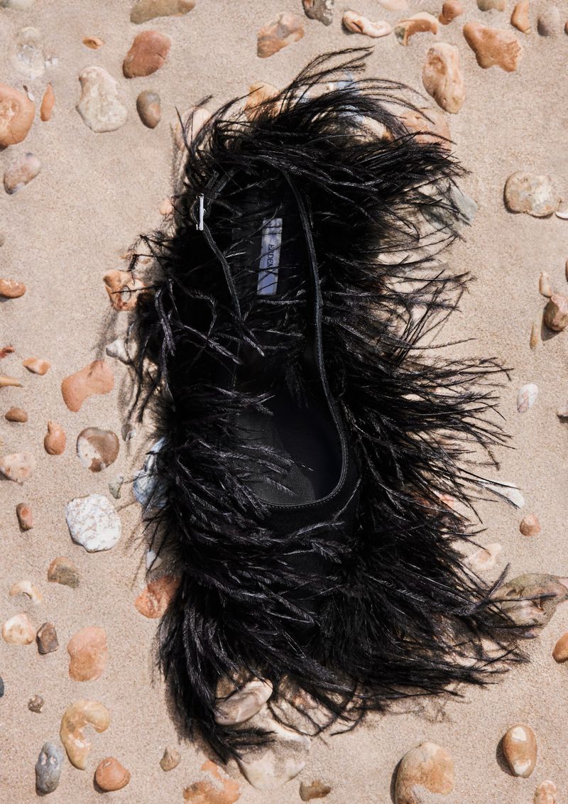 Black feather handbag on a sandy beach with scattered pebbles, showcasing fashion accessory and beach setting in natural lighting.
