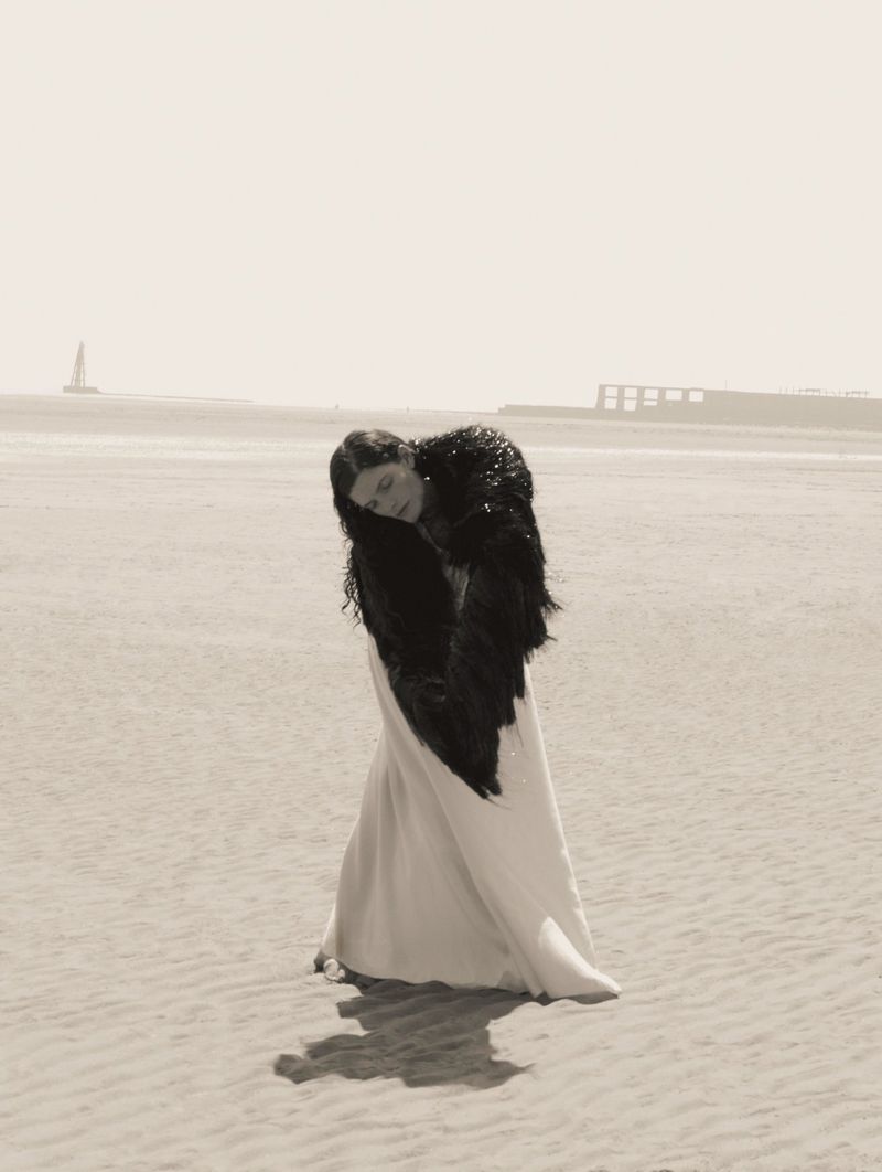 Woman in flowing gown and feathered shawl standing on sandy beach with sail boat in distance under overcast light. Artistic fashion portrait.