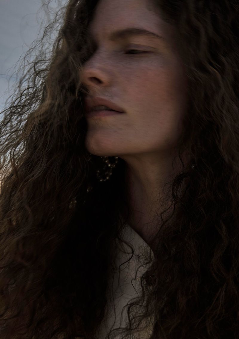 Portrait of a naturally beautiful, light-skinned woman with curly brunette hair, wearing a beige top, against a soft outdoor background.