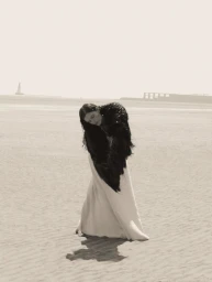 Model in a black textured stole and long white dress standing on a sandy beach under an overcast sky.