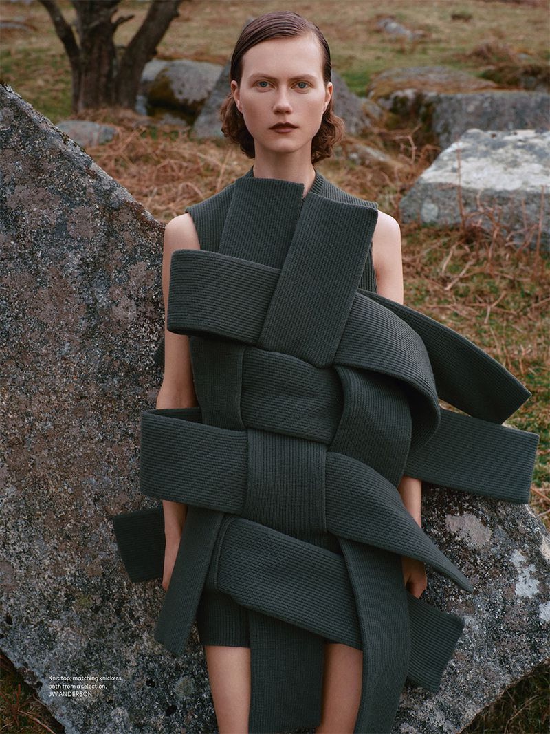 Fashion shot of a fair-skinned woman wearing a dark green, textured, woven garment, standing in a grassy field with large rocks.