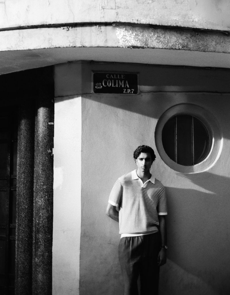 Monochrome portrait of a man with dark hair in a striped polo shirt and dark pants standing in front of a building with a sign.