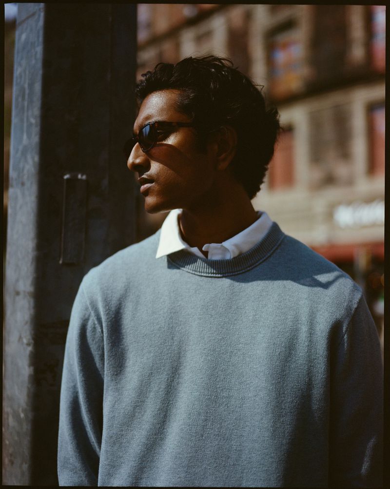 Portrait of a young adult male with dark hair and sunglasses, wearing a blue sweater with a white collared shirt underneath, outdoors.
