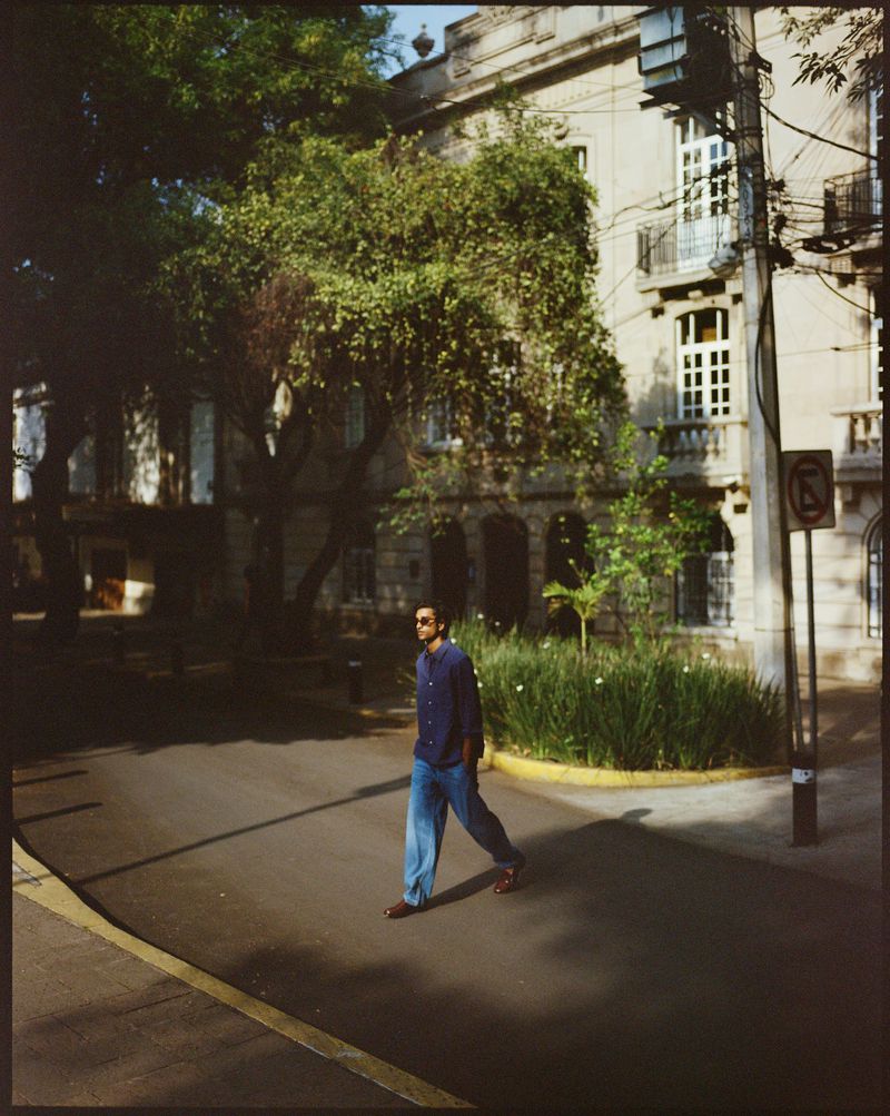 Adult male in sunglasses and casual clothing walks across a crosswalk on a sunny city street with buildings and trees in the background.