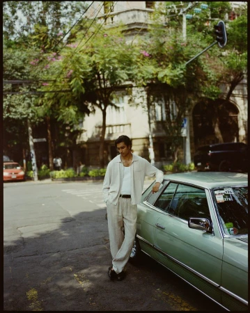 Adult male in casual attire stands by a vintage car on a city street with lush trees and a building in the background on a sunny day.