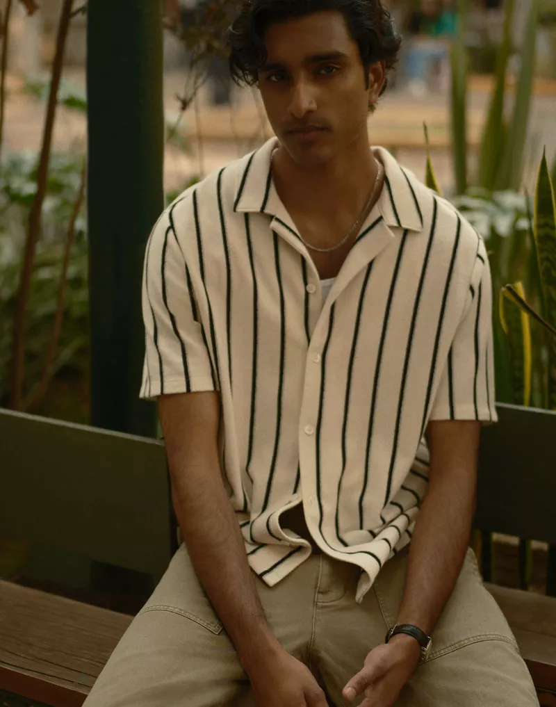 Adult male in vertically striped collared shirt and light brown pants sits on a bench in a verdant urban outdoor setting.