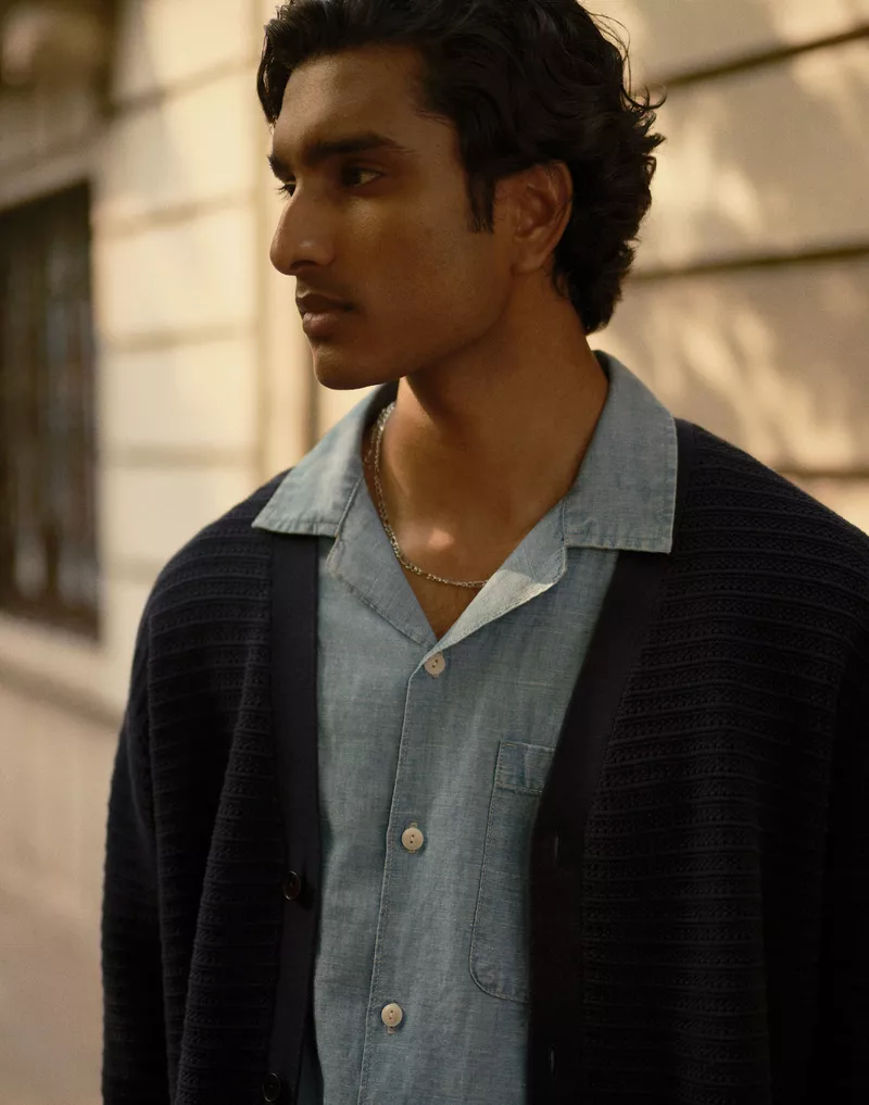 Close-up of a young man with dark hair wearing a light blue button-down shirt and dark cardigan, looking off to the left side.