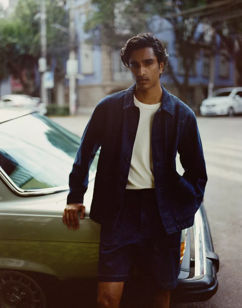 A young man in a blue jacket and shorts leans against a green classic car on a city street with buildings and parked cars in the background.